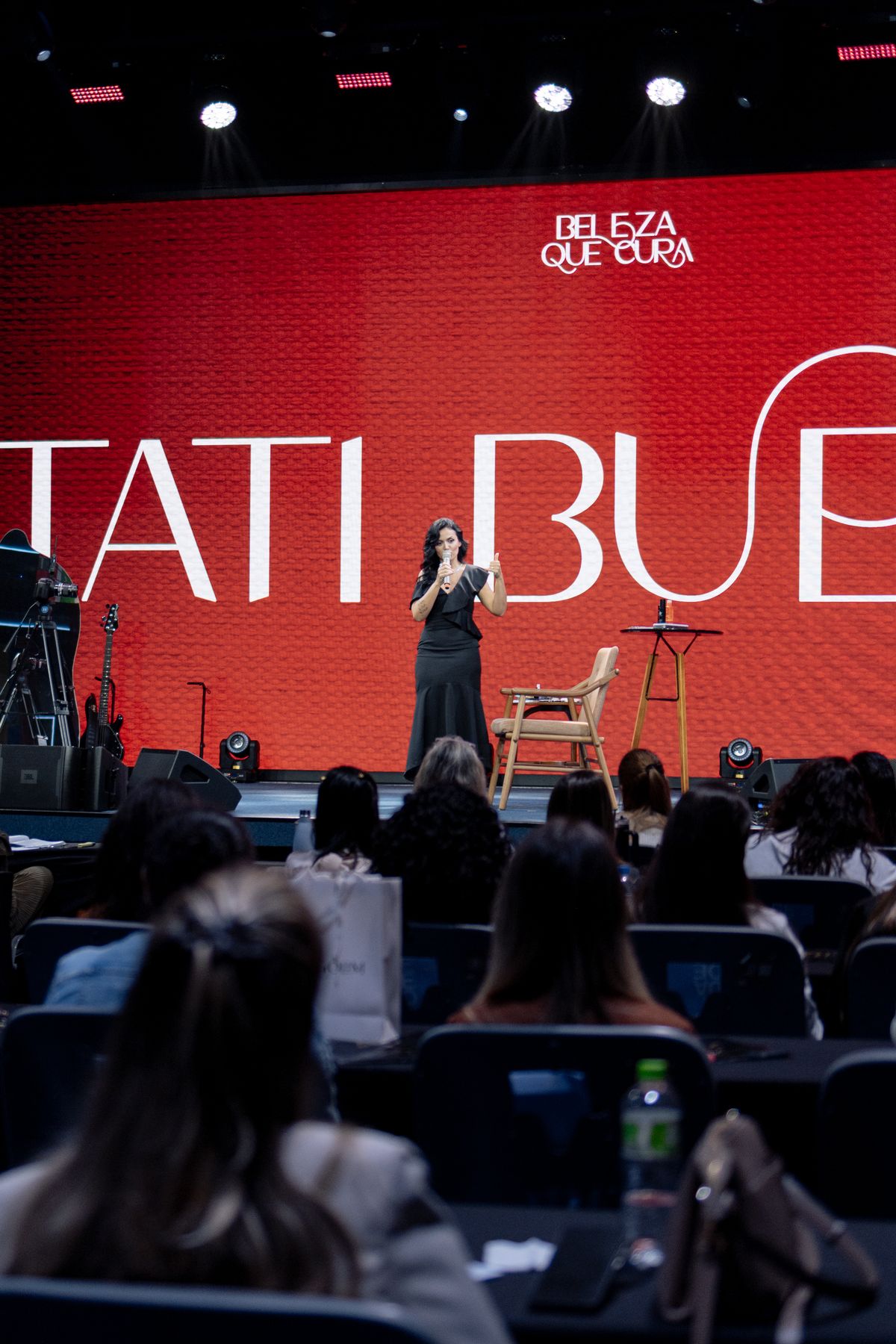 Tati Bueno no palco do Beleza que Cura, diante do telão vermelho com seu nome, com a plateia de mulheres em primeiro plano
